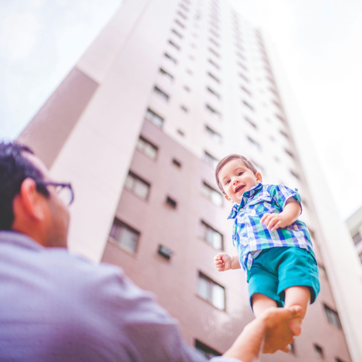 Ein kleiner Junge steht auf der ausgestreckten Hand eines Mannes und wird so in die Höhe gehalten und lacht dabei begeistert. Das Bild ist von unten aufgenommen, im Hintergrund ragen Wolkenkratzer in die Höhe.