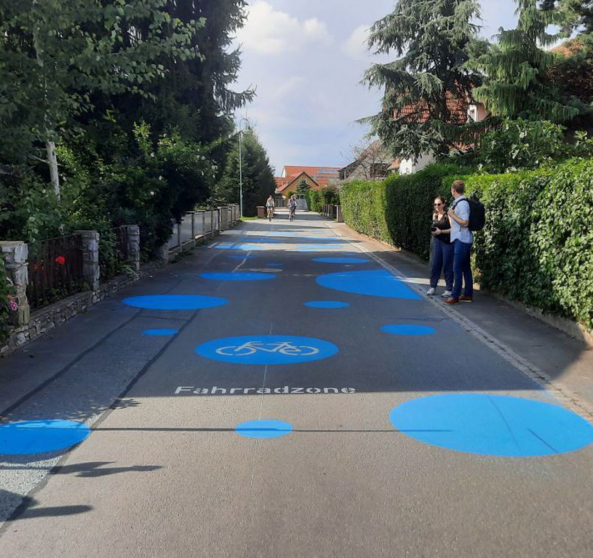 Der Asphaltboden in einer Siedlungsstraße ist mit großen blauen Kreisen als Fahrradzone gekennzeichnet. Am Gehsteig sieht man Fußgänger.
