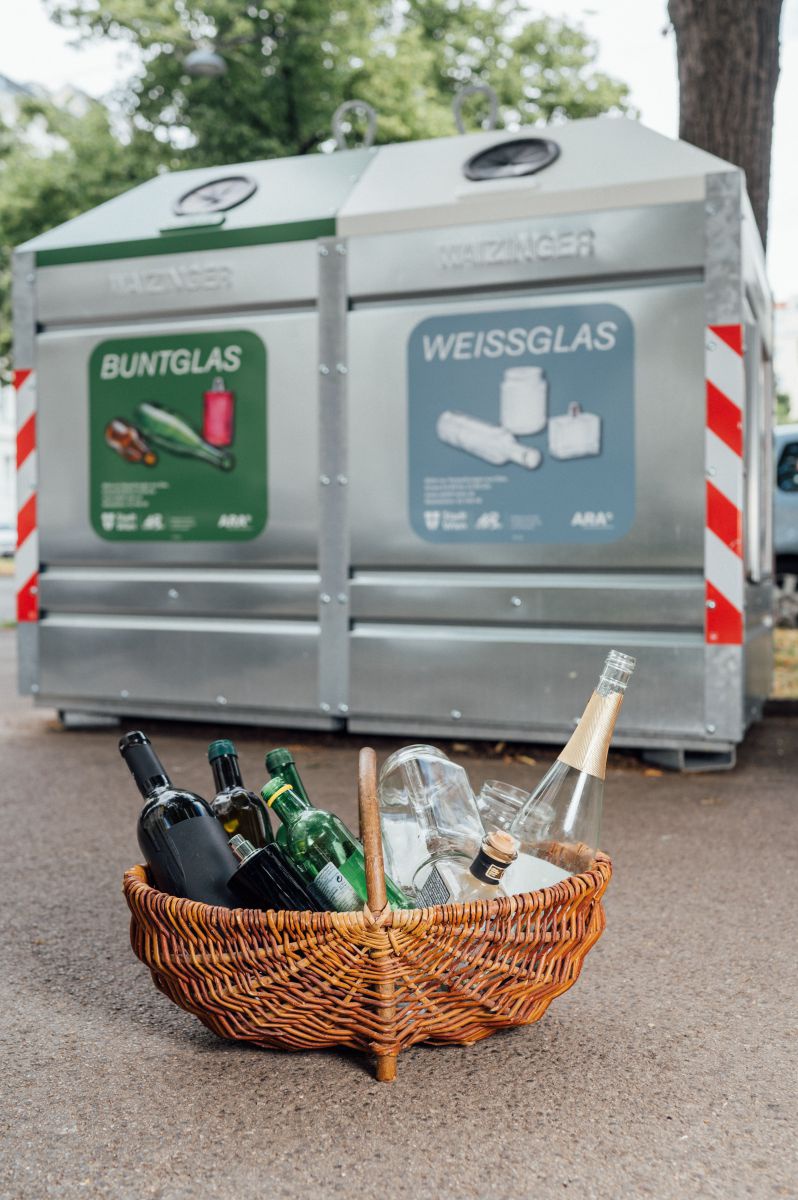 Im Fokus ein Einkaufskorb voller Leerglas, links vom Tragegriff das Buntglas, rechts davon das Weissglas. Im Hintergrund etwas verschwommen der Sammelcontainer.