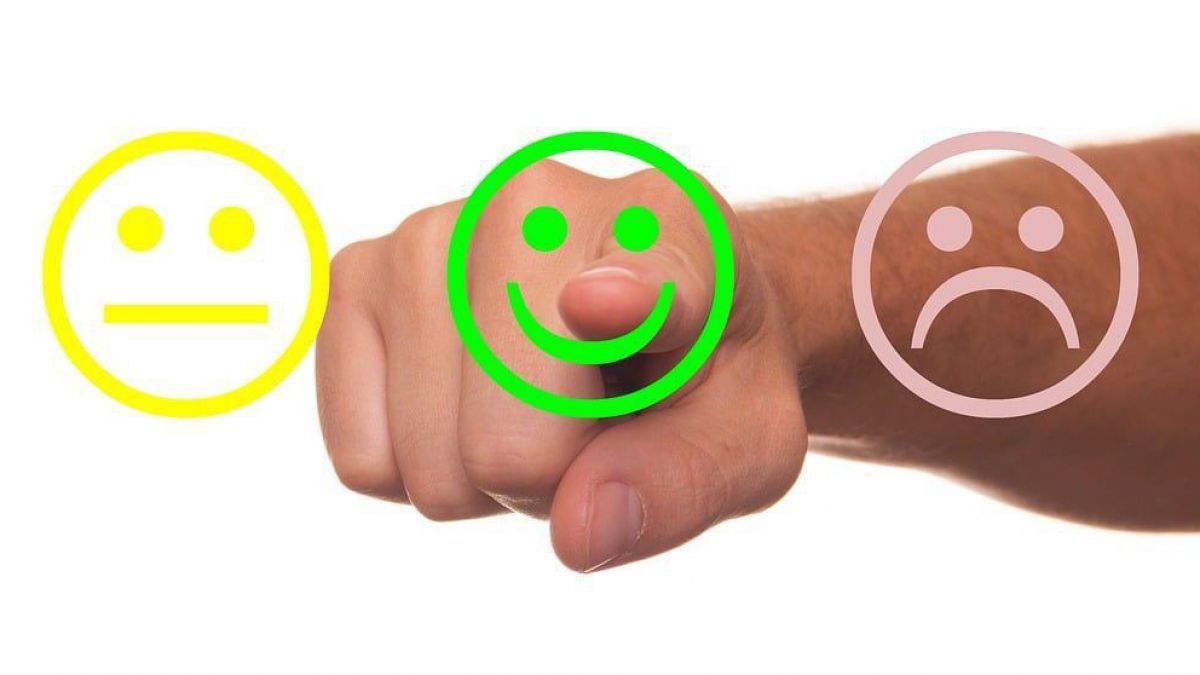 Drei Smileys stehen zur Auswahl: links ein gelber mit geradlinigem Mund, in der Mitte ein grüner mit lachendem Mund und rechts ein grauer mit nach untern gebogenen Mund. Ein Arm kommt aus dem Hintergrund und tippt mit dem Zeigefinger auf den lachenden Smi