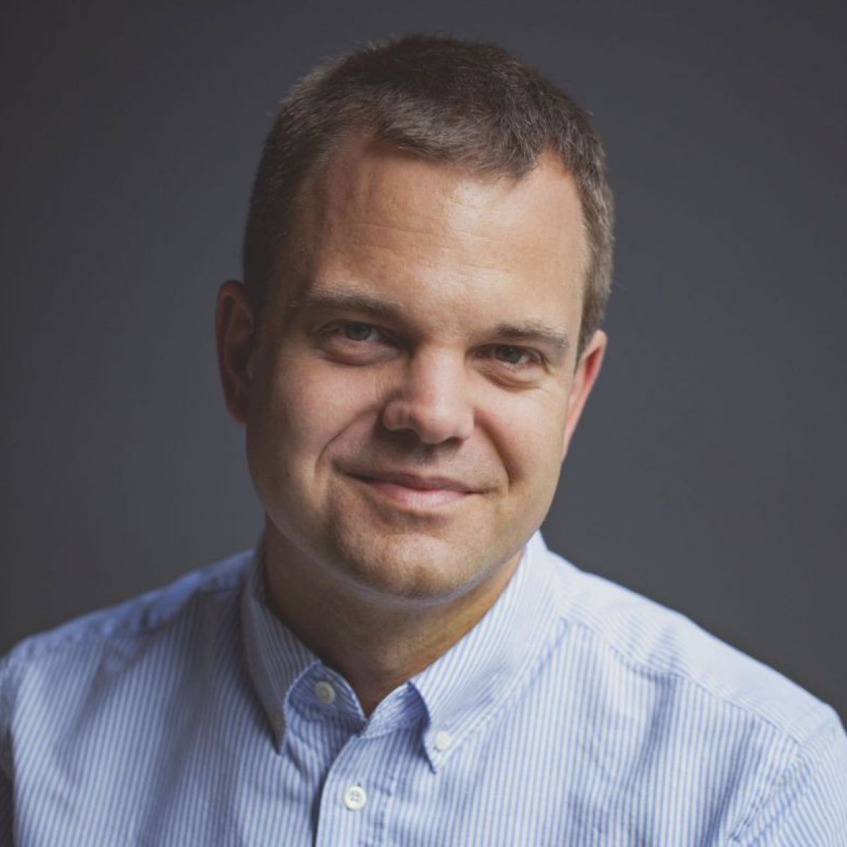 Portraitfoto vor dunklelgrauem Hintergrund: Hinnerk Hansen trägt ein fein gestreiftes Hemd in Weiß-Hellblau.